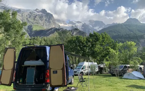 En campingplats med flera husbilar, inklusive en blå med öppna bakdörrar, i en dal med snöklädda berg i bakgrunden. En perfekt plats för äventyr med edins husbilar.