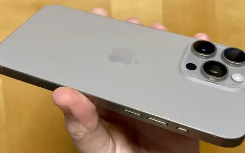 En hand håller en ljusgrå iPhone 15. Kameramodulen på baksidan har tre linser, redo att fånga dina bästa ögonblick.