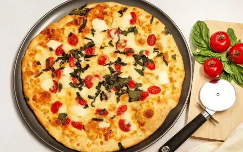 Pyszna pizza z serka wiejskiego, pomidorkami koktajlowymi i bazylią. Obok leżą świeże pomidory i krajalnica do pizzy.