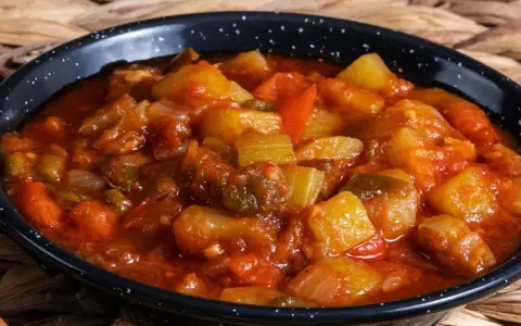 Un plato humeante de pisto manchego, con trozos de calabacín, pimiento y tomate en una salsa rojiza, servido en una cazuela negra.