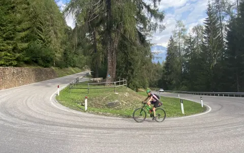 Ciclista su strada tortuosa, immersa nel verde del **campo Carlo Magno**, con alberi secolari e montagne sullo sfondo.