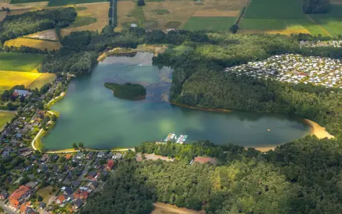 Luftaufnahme des Feldmarksees Sassenberg mit einer kleinen Insel, Booten am Steg, einem Wohngebiet und Feldern.