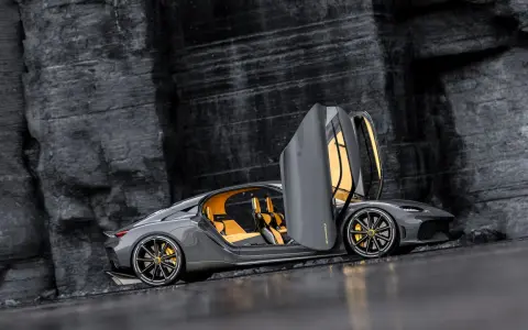 Szary Koenigsegg Gemera, czteromiejscny supersamochód z otwartymi drzwiami, prezentuje swoje żółte wnętrze na tle skalistej ściany.