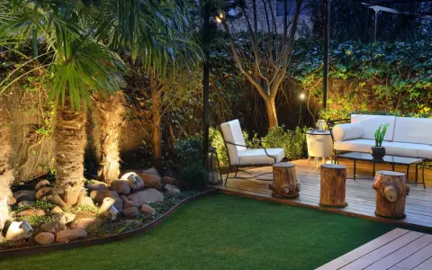 Un jardín poco mantenimiento con césped artificial, palmeras iluminadas y una zona lounge con sofá blanco y sillón.
