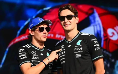 Dos pilotos de Fórmula 1, George Russell y un compañero, sonríen con gafas de sol. Russell lleva una gorra azul y ambos visten camisetas negras con logotipos de patrocinadores.