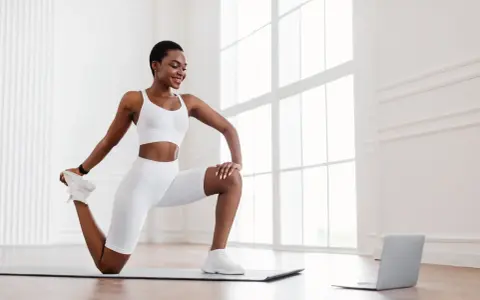 Femme en tenue de sport blanche effectuant un exercice quadriceps sur un tapis, guidée par un ordinateur portable.
