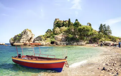 Ein Boot liegt im kristallklaren Wasser vor einer felsigen Insel mit üppiger Vegetation. Das sizilien wetter im oktober ist perfekt für einen entspannten Tag am Strand.