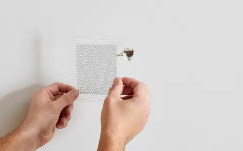 Hands apply a mesh patch to a hole in a white wall, demonstrating how to patch a hole in the wall.
