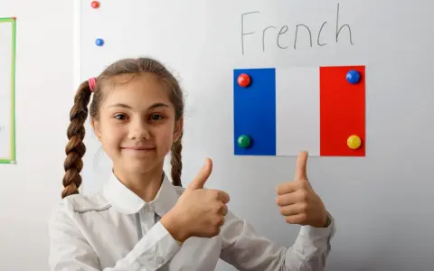 Uśmiechnięta dziewczynka z warkoczami pokazuje kciuki w górę na tle tablicy z napisem "French" i flagą Francji. To symbol pozytywnego podejścia do nauki, być może w ramach systemu edukacji we Francji.