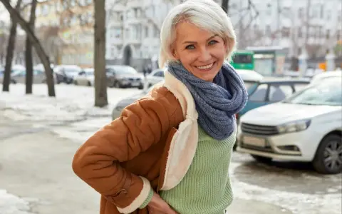 Uśmiechnięta kobieta w brązowym, sztucznym kożuszku damskim i niebieskim szaliku pozuje na tle zimowego miasta.