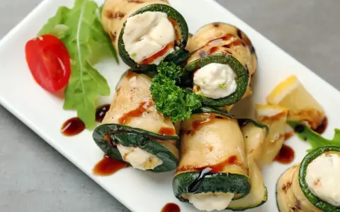 Deliziosi involtini di zucchine grigliate ripieni di formaggio cremoso, guarniti con pomodorini, rucola e salsa scura.