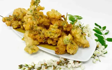 Fiori di acacia fritti, dorati e croccanti, serviti su un piatto bianco con rametti di fiori e foglie fresche.
