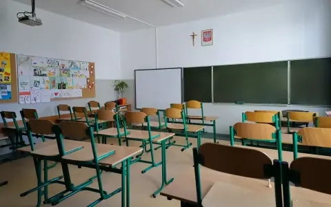 Pusta klasa z rzędami ławek i krzeseł, tablicą i dziecięcymi rysunkami na ścianie. Brak ucznia w tej sali może sugerować nieklasyfikowanie ucznia.