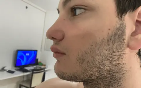 Perfil de un joven con barba incipiente, preparándose para afeitar barba. Al fondo, un monitor con pantalla azul.