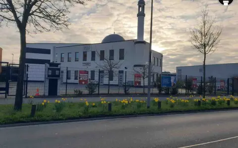 Moderne Subhan Moschee mit Minarett und Kuppel, umgeben von einem Zaun mit Schildern. Gelbe Narzissen blühen davor.