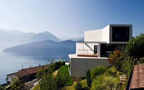 Modernes Haus in Hanglage mit Blick auf einen See und Berge. Ein traditionelles Haus liegt tiefer am Hang.