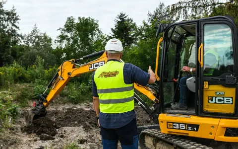 Szkolenie na koparkę w Akademii Operatora. Instruktor w kamizelce odblaskowej pokazuje, jak uzyskać uprawnienia na koparkę.