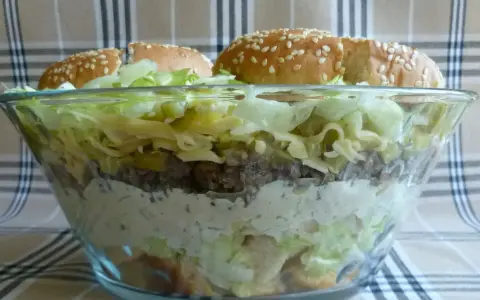 Warstwowa sałatka big mac w szklanej misie, z bułkami na wierzchu.