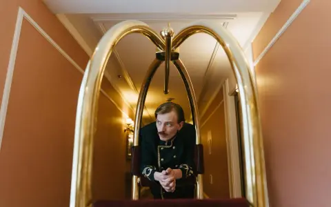 Elegancki hotelarz w historycznym mundurze czeka przy wózku bagażowym w hotelowym korytarzu.