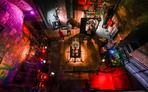 W muzeum nauki gdańskiej zwiedzający podziwiają mechanizmy i eksponaty. Kolorowe światła podkreślają atmosferę.