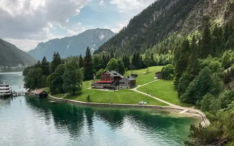 Gaisalm am Achensee: ein Gasthaus mit roten Fensterläden, umgeben von grünen Wiesen und Bergen. Ein Schiff liegt am Steg.