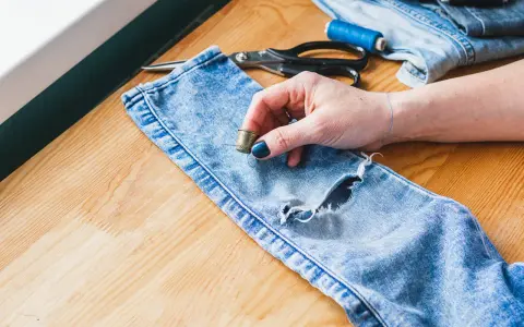 Ręka z naparstkiem i igłą przygotowuje się, by pokazać, jak zacerować dziurę w jeansach. Nożyczki i nici leżą obok.