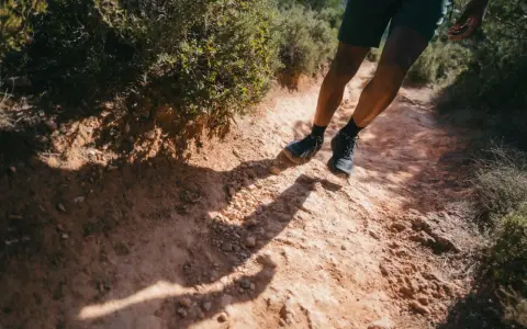 Jambes d'un homme en short et chaussures de trail, s'engageant sur un sentier rocailleux et poussiéreux. L'ombre de son corps s'étire sur le chemin, suggérant un rythme de fast hiking sous le soleil.