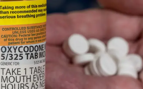 Zbliżenie na butelkę z lekiem Oxycodone i garść białych tabletek w dłoni.