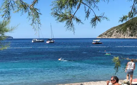 En man sitter på en stenstrand vid havet. I vattnet simmar en person och flera båtar ligger för ankar. En grön kulle reser sig i bakgrunden. En idyllisk vy från Vis, Kroatien.