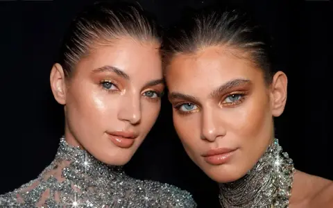Dos mujeres con maquillaje dorado y un sutil rabillo del ojo, luciendo collares brillantes.