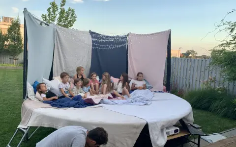 Dzieci siedzą na wielkim materacu pod gołym niebem, oglądając film. To idealne urodziny na dworze z magicznymi światełkami i wygodnymi kocami.