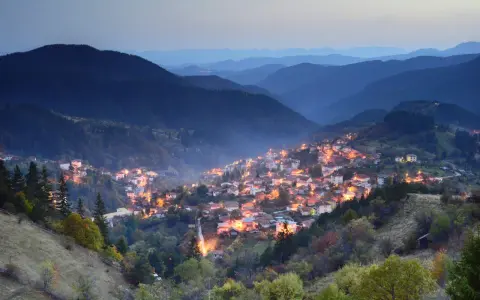Wieczorna panorama górskiej wioski w Bułgarii. Domy rozświetlone ciepłym światłem, otoczone przez ciemne, zalesione wzgórza.