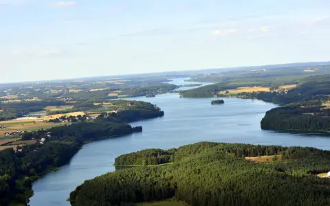 Rozległe jezioro raduńskie dolne otoczone lasami i polami. W oddali widać zabudowania.