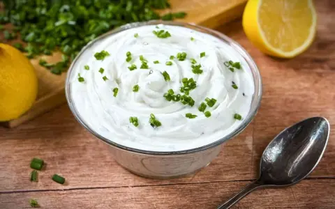 Cremige Sour Cream, garniert mit Schnittlauch, neben Zitronenspalten und einem Löffel auf Holzuntergrund.