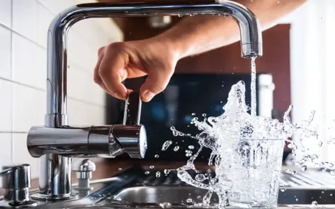 Mano girando la llave para cuidar el agua, que salpica al llenar un vaso.