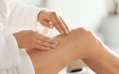Kobieta w białym szlafroku wykonuje peeling nóg. To idealny peeling przed goleniem, by skóra była gładka.