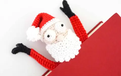 Marcador de libro amigurumi de Papá Noel, con brazos rojos y gorro festivo, asomando de un libro rojo.