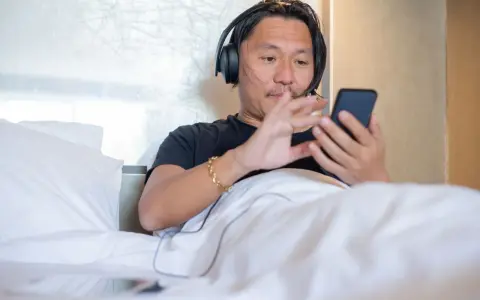 Mężczyzna w słuchawkach leży w łóżku, korzystając z telefonu. To prosty sposób, jak słuchać audiobooka na telefonie, relaksując się.