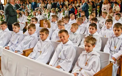 Dzieci w bieli, gotowe na rocznicę komunii świętej, siedzą w kościele. W tle widać zgromadzonych gości i fotografa.