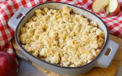 Ein goldbrauner Apfel Crumble in einer Auflaufform, bereit zum Genießen. Daneben liegen frische Äpfel auf kariertem Tuch.