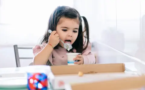 Mała dziewczynka z kitkami je jogurt z kubeczka. Od kiedy dziecko może jeść danonki? Odpowiedź jest prosta: gdy tylko zaczyna jeść pokarmy stałe.