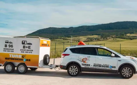 Un coche blanco de **4x4 autoescuela** con remolque, listo para enseñar a conducir en un paisaje rural.