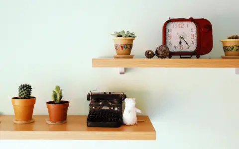 Ideas para decorar baldas salón: dos estantes de madera con cactus, una máquina de escribir antigua, un peluche blanco y un reloj rojo.