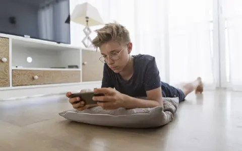 Un adolescent, absorbé par son téléphone, illustre la dépendance aux écrans. Il est allongé sur un coussin, le regard fixé sur l'appareil.