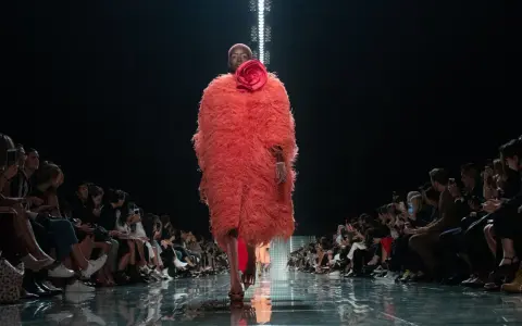 Modella con cappotto vaporoso color corallo e fiore rosso, sfila su una passerella lucida. Il pubblico ammira gli abbinamenti audaci.