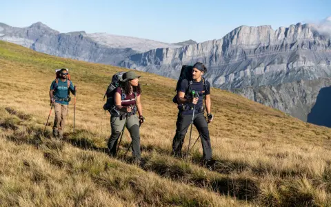 Trzy osoby z plecakami i kijkami trekkingowymi idące po trawiastym zboczu górskim.