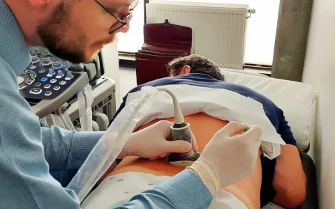 Lekarz wykonuje zabieg inwazyjny, co to znaczy? Używa aparatu USG do badania pacjenta leżącego na kozetce.