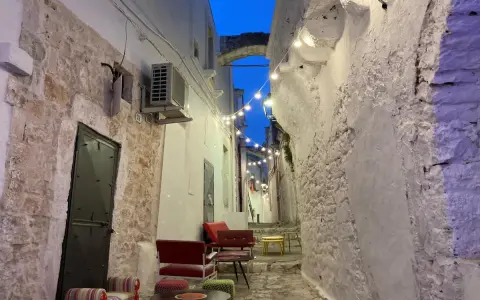 Un vicolo suggestivo nel Val d'Itria, illuminato da lucine, con sedute colorate e muri in pietra bianca.