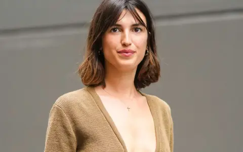 Mujer con cabello castaño ondulado, en transición de pelo corto a largo, luce un suéter marrón y un collar con cruz.