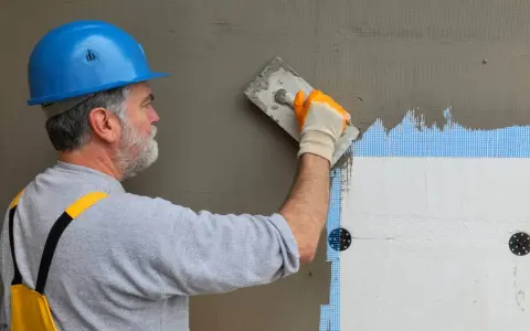 Budowlaniec w niebieskim kasku nakłada tynk na styropian. Prace nad fasadą, w tym suche tynki, postępują.